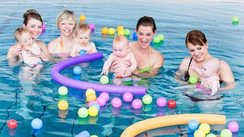 Vier Frauen stehen im Wasser und halten Babys, umgeben von bunten Bällen und Schaumstoffringen in einem Schwimmbad.